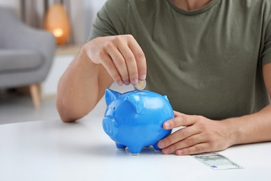 Photo of Young man putting coin into piggy bank at table, closeup