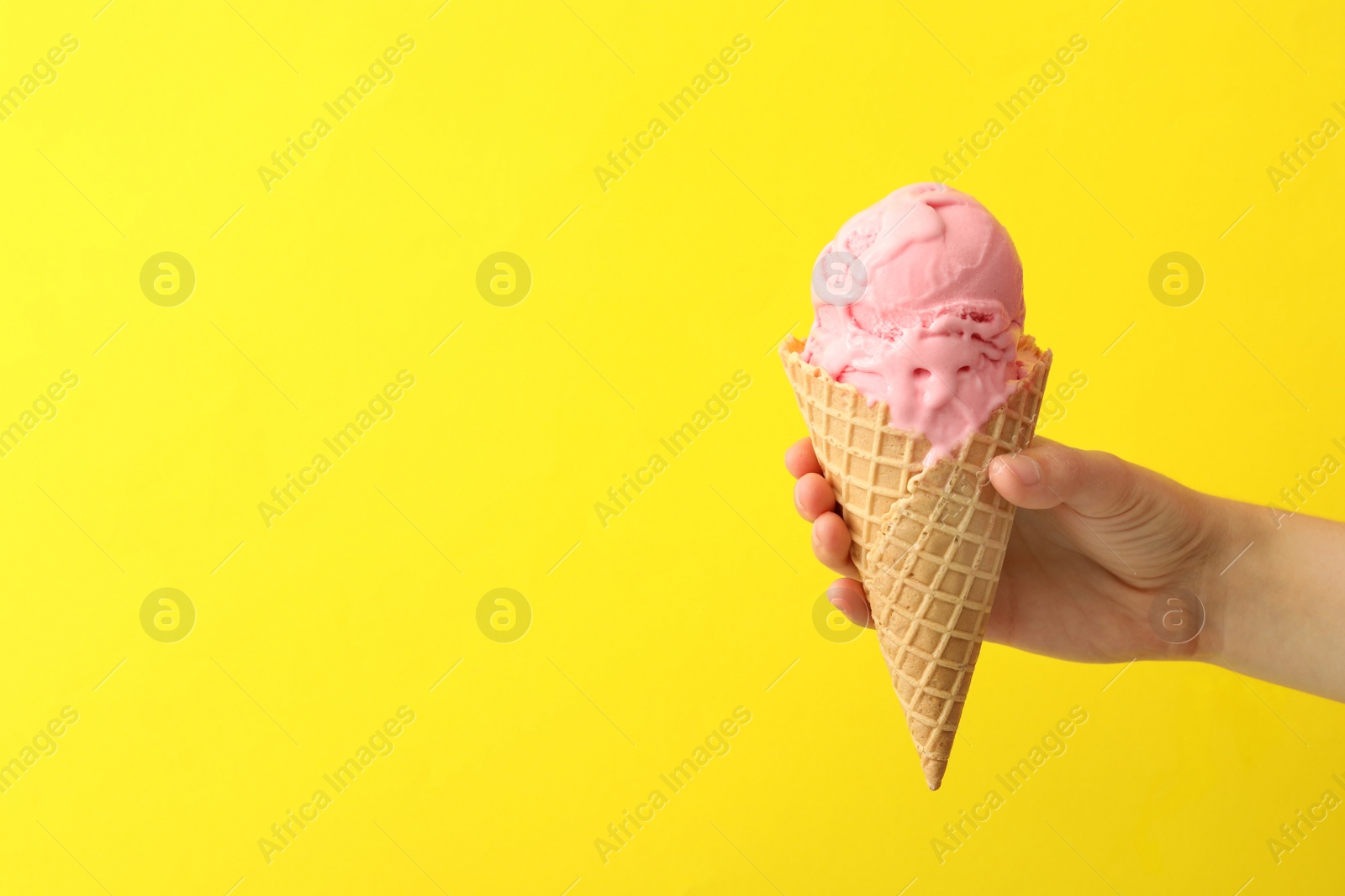 Photo of Woman holding waffle cone with delicious pink ice cream on yellow background, closeup. Space for text