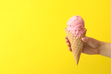 Woman holding waffle cone with delicious pink ice cream on yellow background, closeup. Space for text