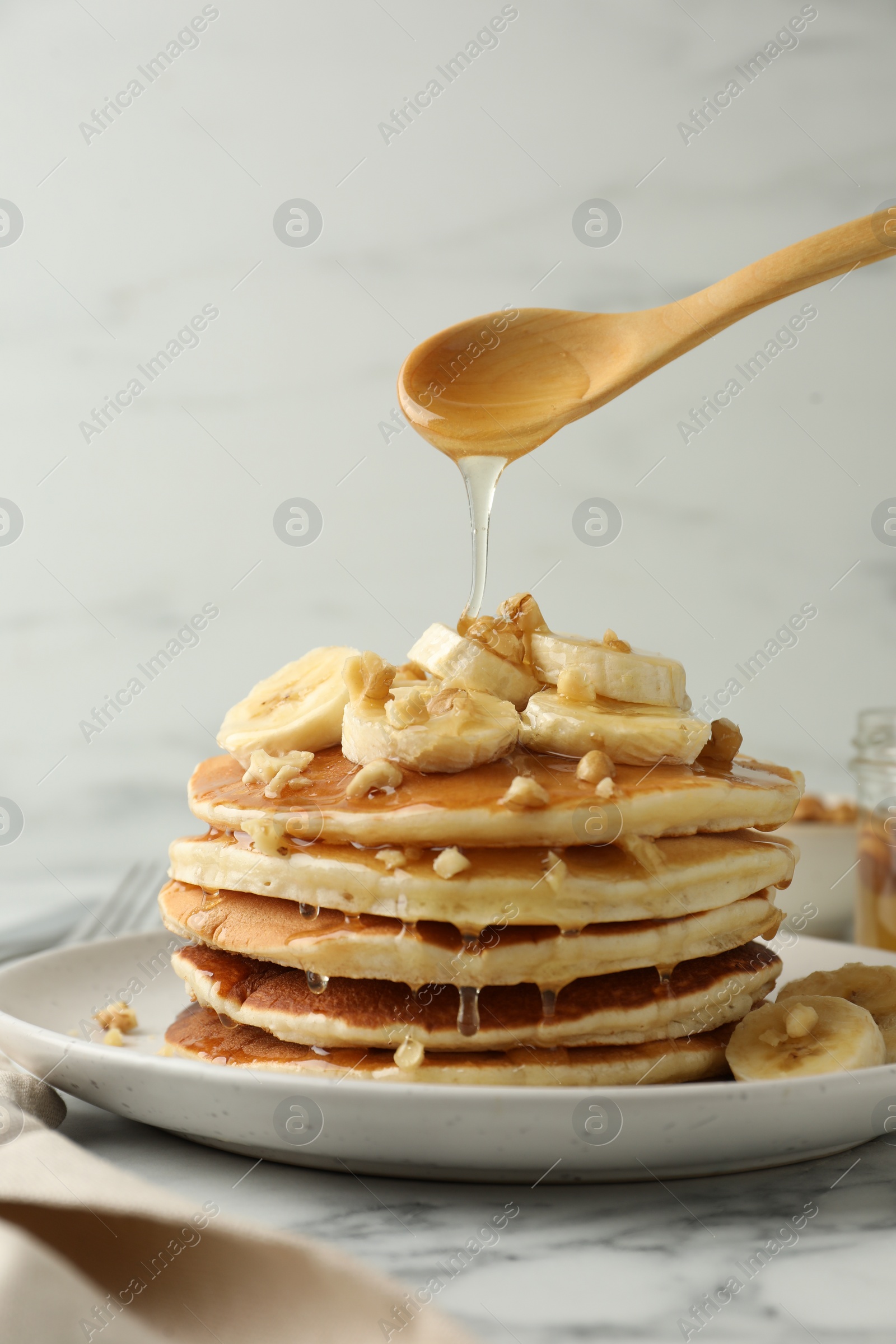 Photo of Pouring honey from spoon onto delicious pancakes with bananas and walnuts at white marble table