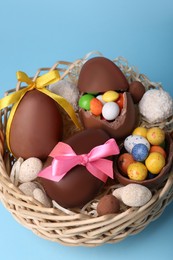 Wicker basket with tasty chocolate Easter eggs and different candies on light blue background