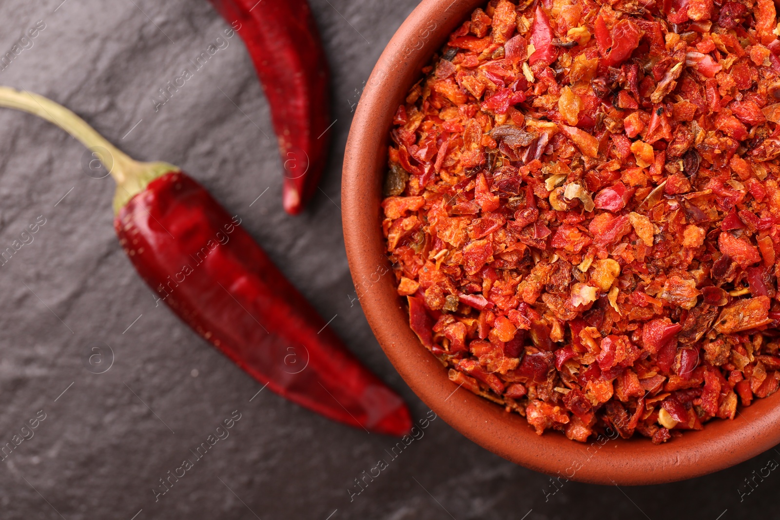 Photo of Chili pepper flakes in bowl and pods on dark textured table, flat lay