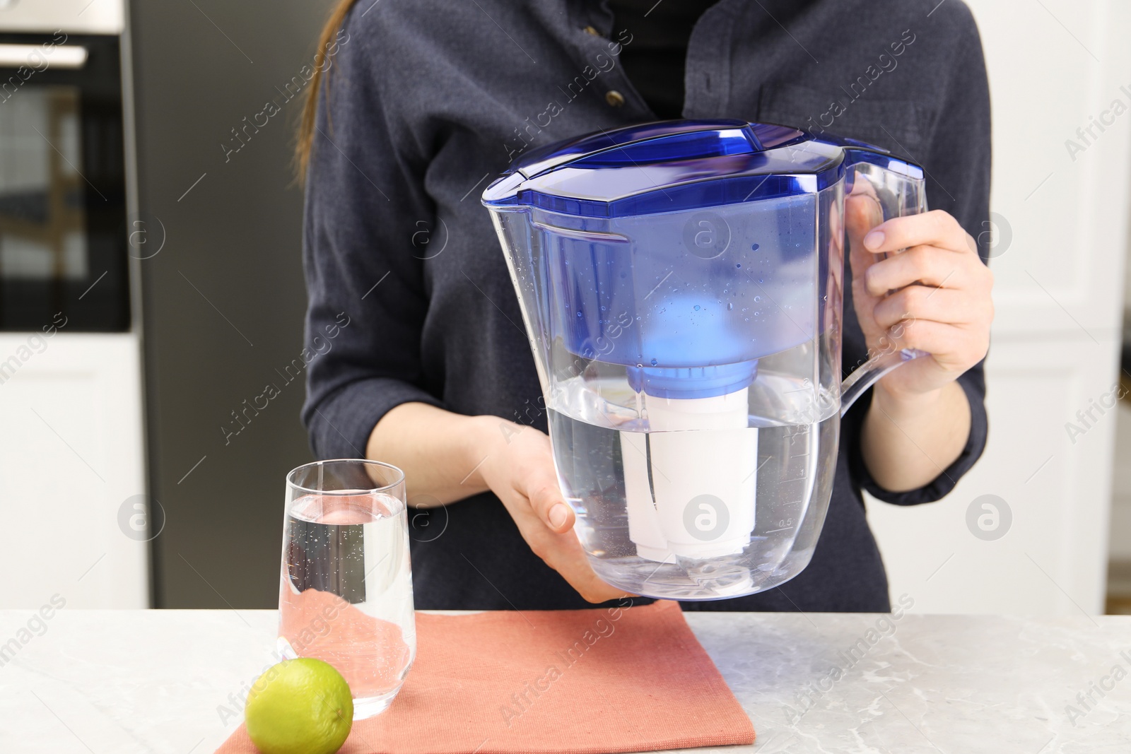 Photo of Woman with water filter jug at light marble table in kitchen, closeup