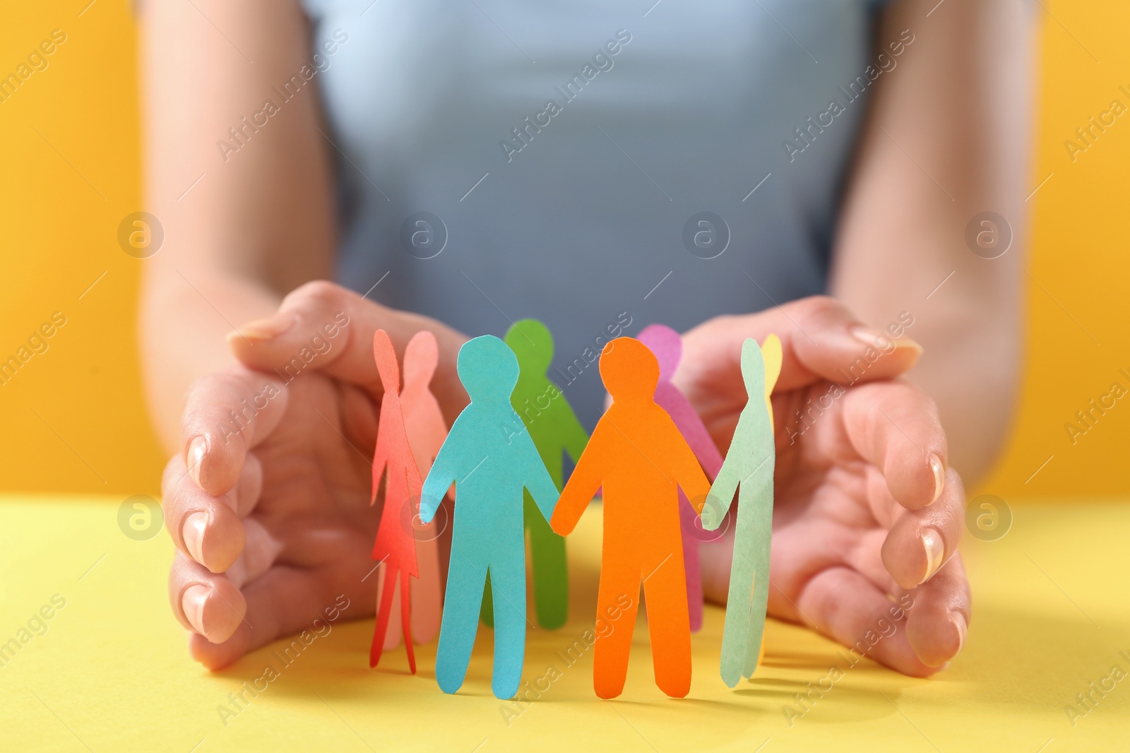 Photo of Woman protecting paper human figures on yellow table, closeup. Diversity and inclusion concept