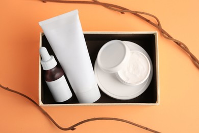 Box with cosmetic products and decorative twigs on orange background, flat lay