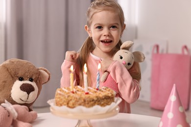 Cute girl with birthday cake and toys at table indoors