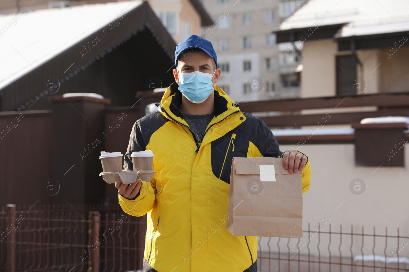 Photo of Courier in medical mask holding takeaway food and drinks near house outdoors. Delivery service during quarantine due to Covid-19 outbreak