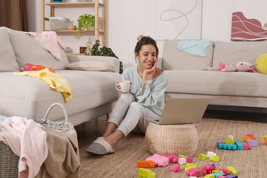 Photo of Young mother drinking tea and working with laptop on floor in messy room