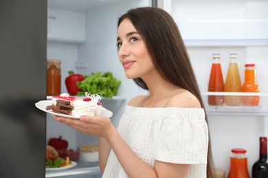 Photo of Concept of choice between healthy and junk food. Woman holding plate with sweets near refrigerator in kitchen