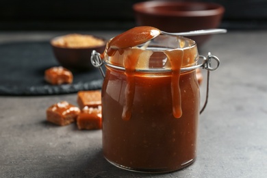 Photo of Jar with tasty caramel sauce and spoon on grey table
