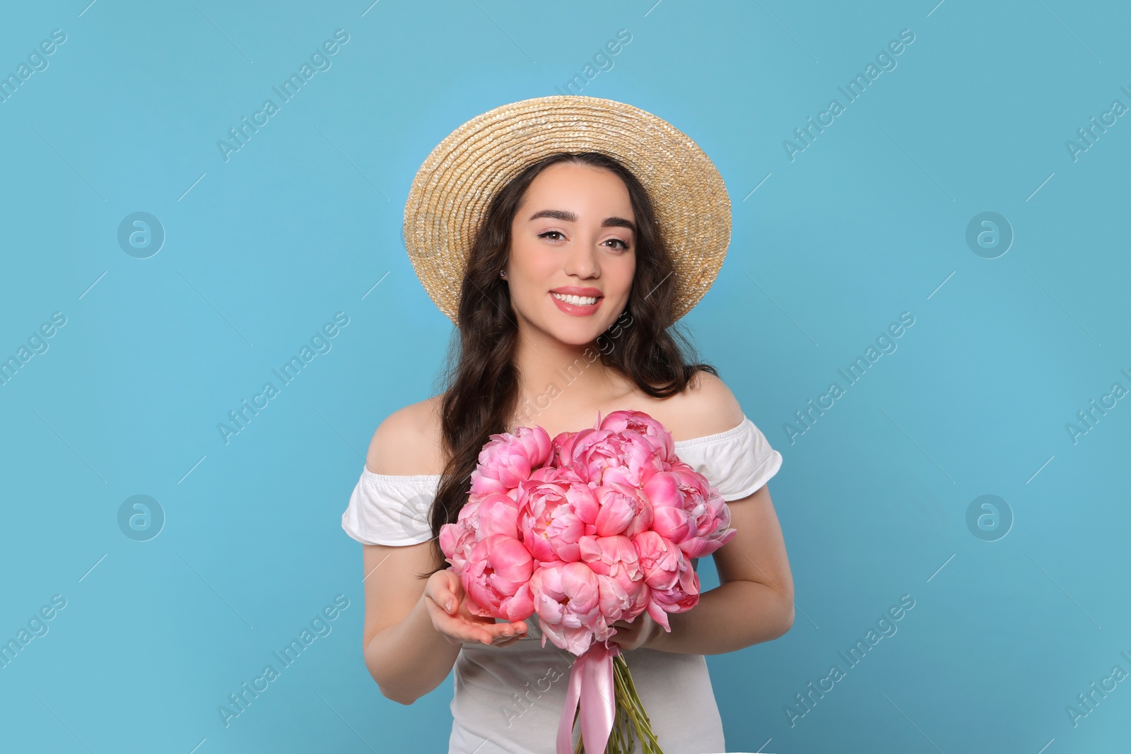 Photo of Beautiful young woman in straw hat with bouquet of pink peonies against light blue background