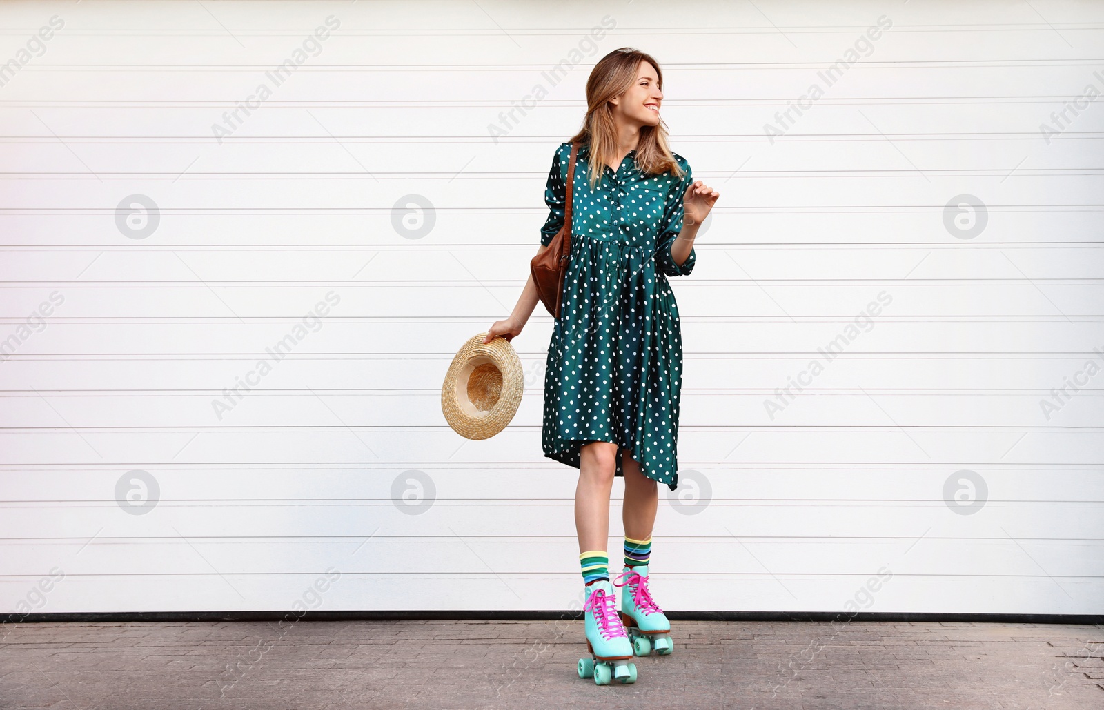 Photo of Happy stylish young woman with vintage roller skates, hat and backpack near white garage door on street