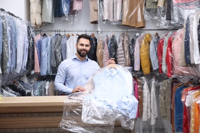 Photo of Dry-cleaning service. Happy worker holding hangers with clothes in plastic bags at counter indoors