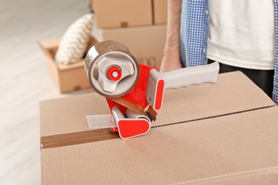 Photo of Moving box and adhesive tape dispenser indoors, closeup