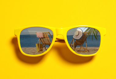 Stylish sunglasses with reflection of sandy shore on yellow background, top view 
