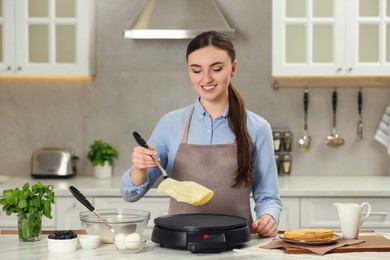 Happy woman cooking delicious crepe on electric maker at white marble table in kitchen