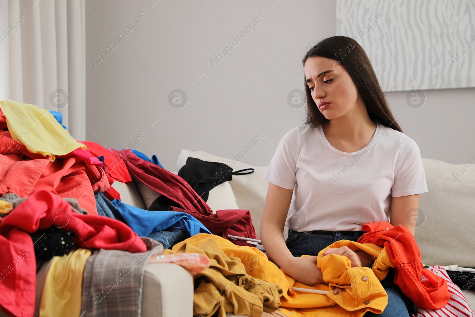 Photo of Upset young woman with lots of clothes on sofa in room. Fast fashion