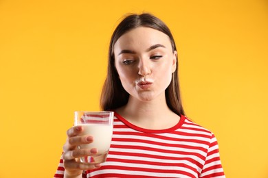 Beautiful woman with milk mustache holding glass of tasty dairy drink on orange background