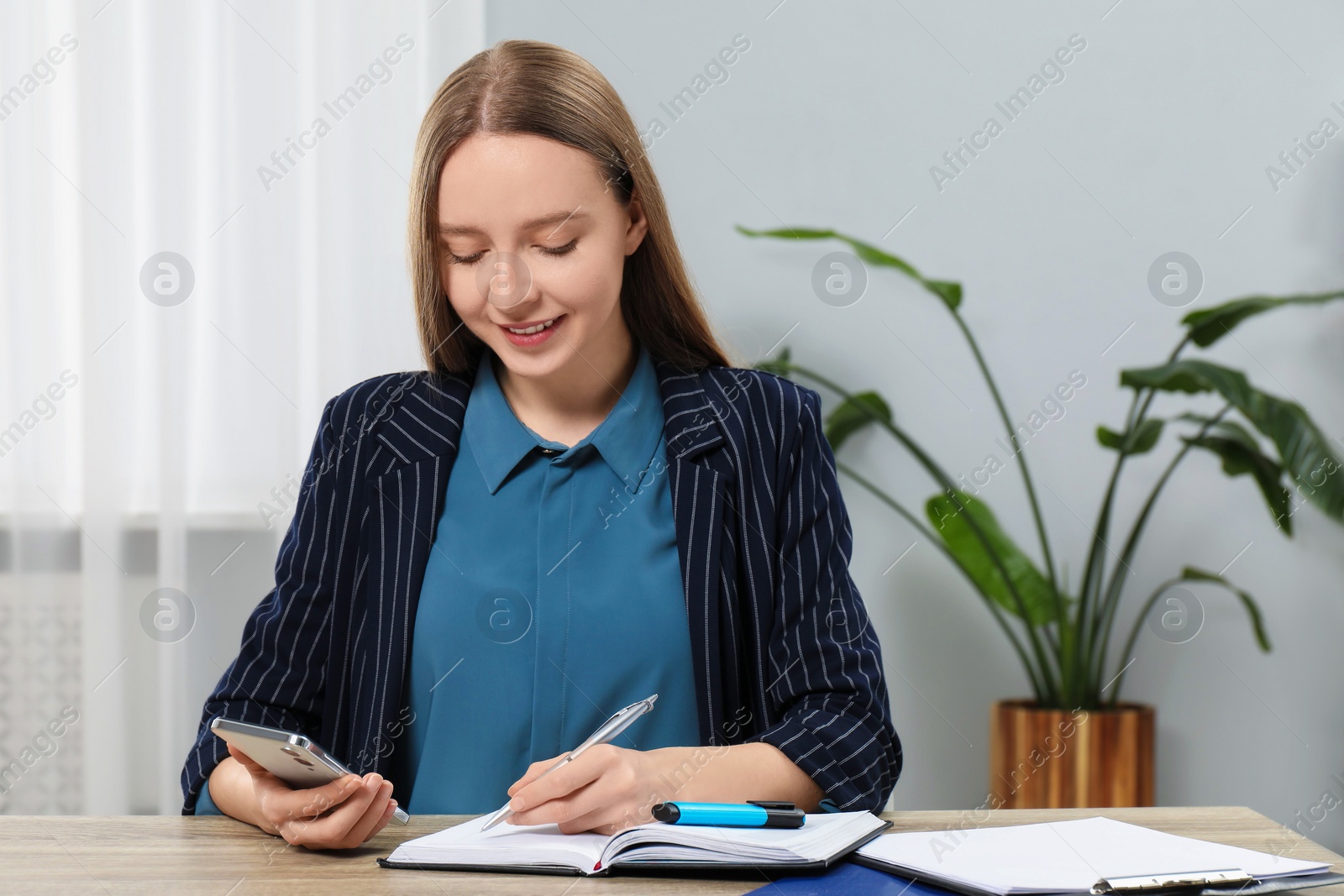 Photo of Woman taking notes while using smartphone at wooden table in office