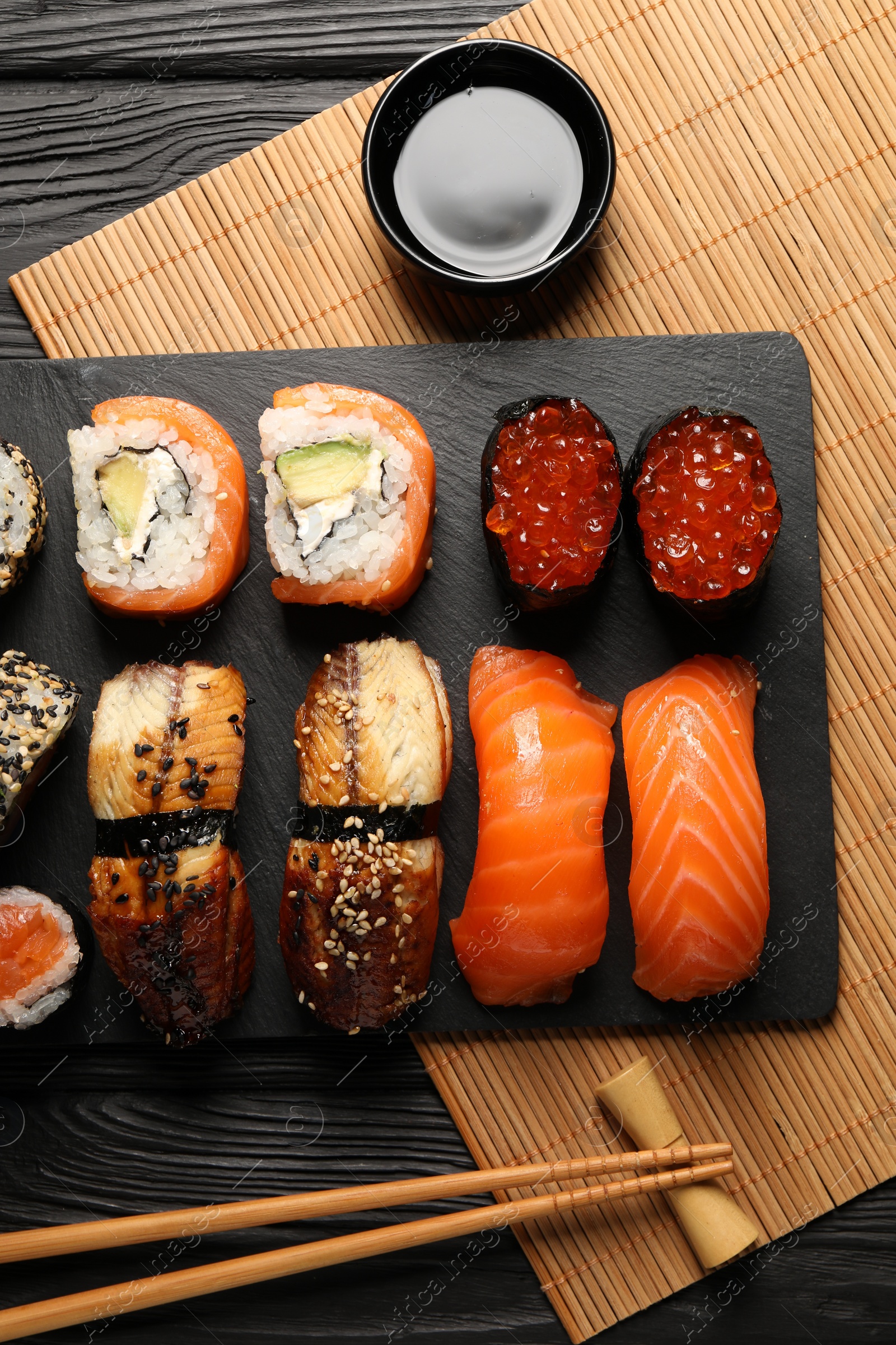 Photo of Delicious sushi rolls, soy sauce and chopsticks on black wooden table, flat lay