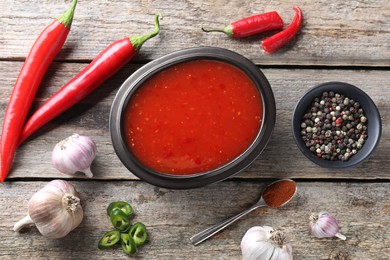 Spicy chili sauce in bowl and ingredients on wooden table, flat lay