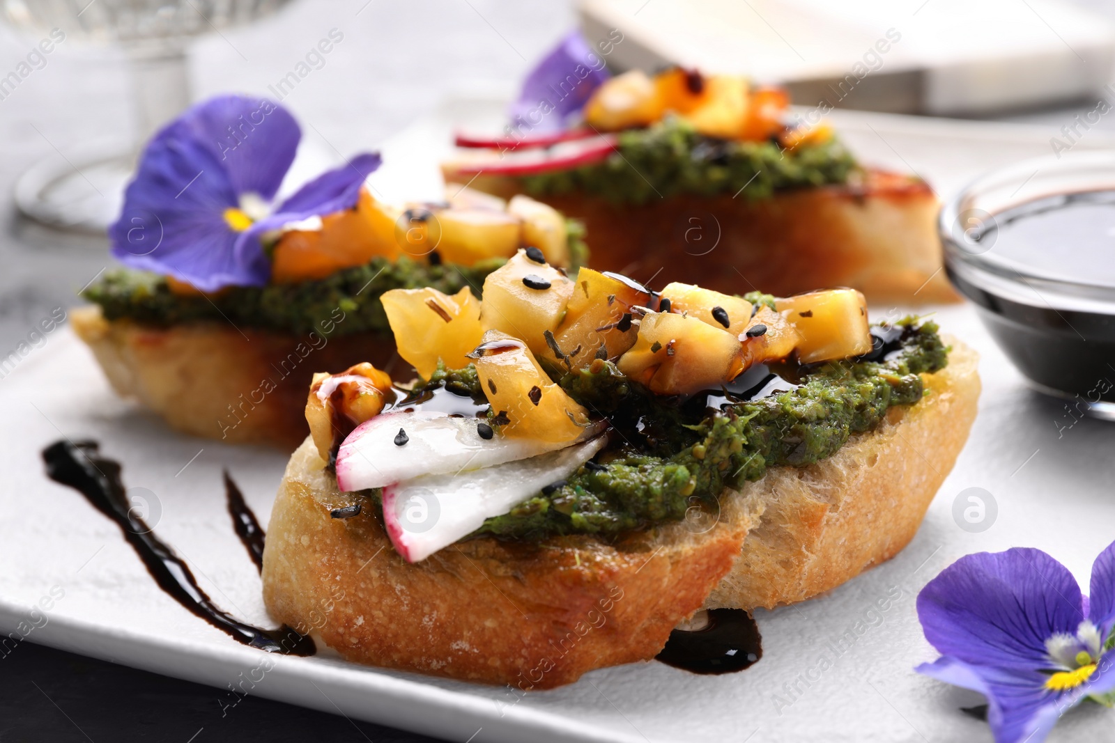 Photo of Delicious bruschettas with pesto sauce, tomatoes, balsamic vinegar and violet flowers on white plate, closeup