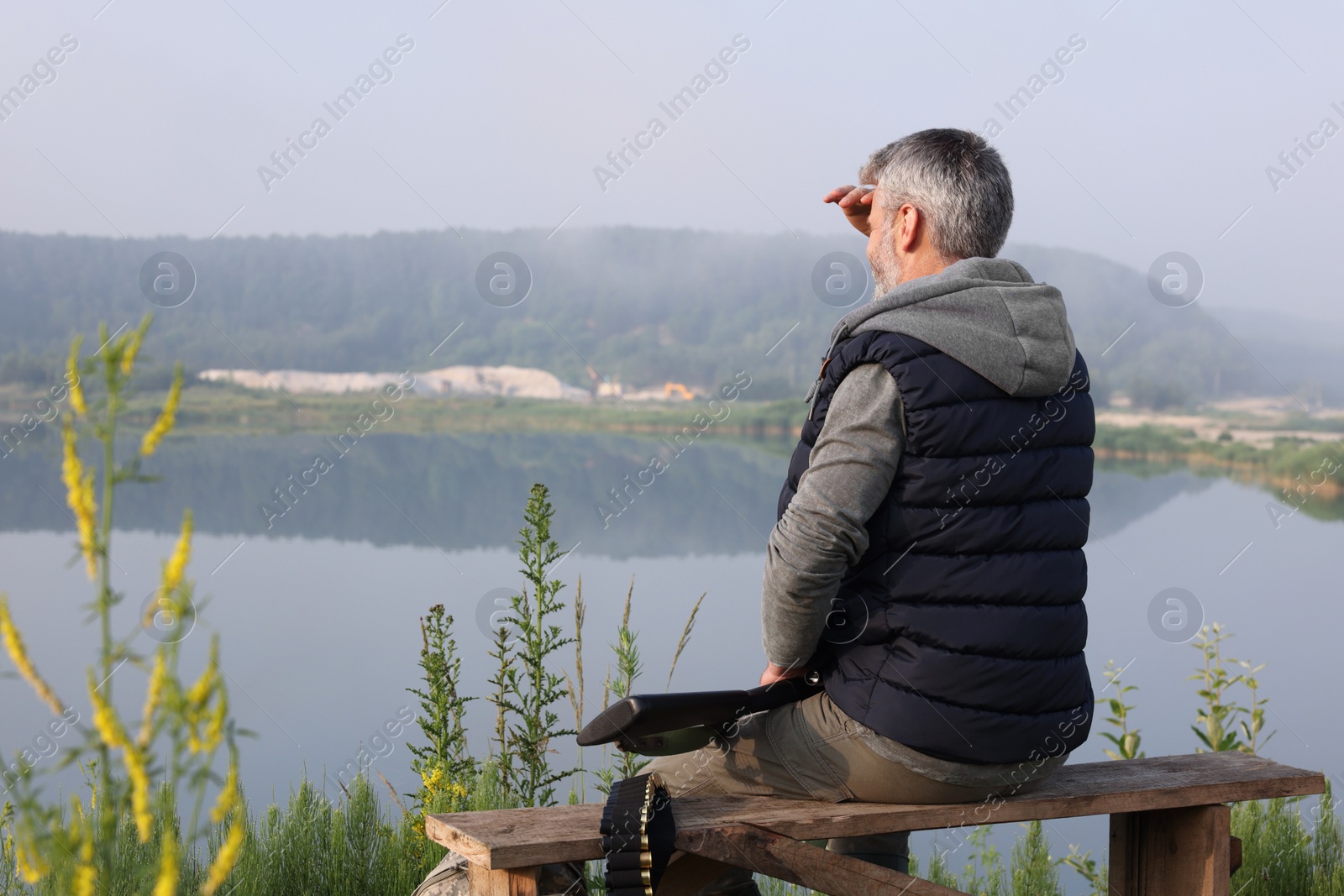 Photo of Man with hunting rifle sitting on wooden bench near lake outdoors, back view. Space for text