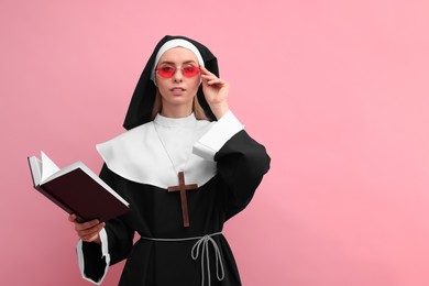 Photo of Woman in nun habit and sunglasses holding Bible against pink background. Space for text