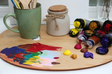 Wooden artist's palette with colorful paints, tubes and scented candle on white table