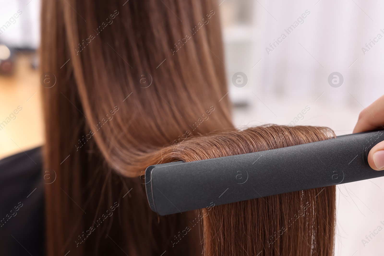 Photo of Hairdresser straightening woman's hair with flat iron indoors, closeup