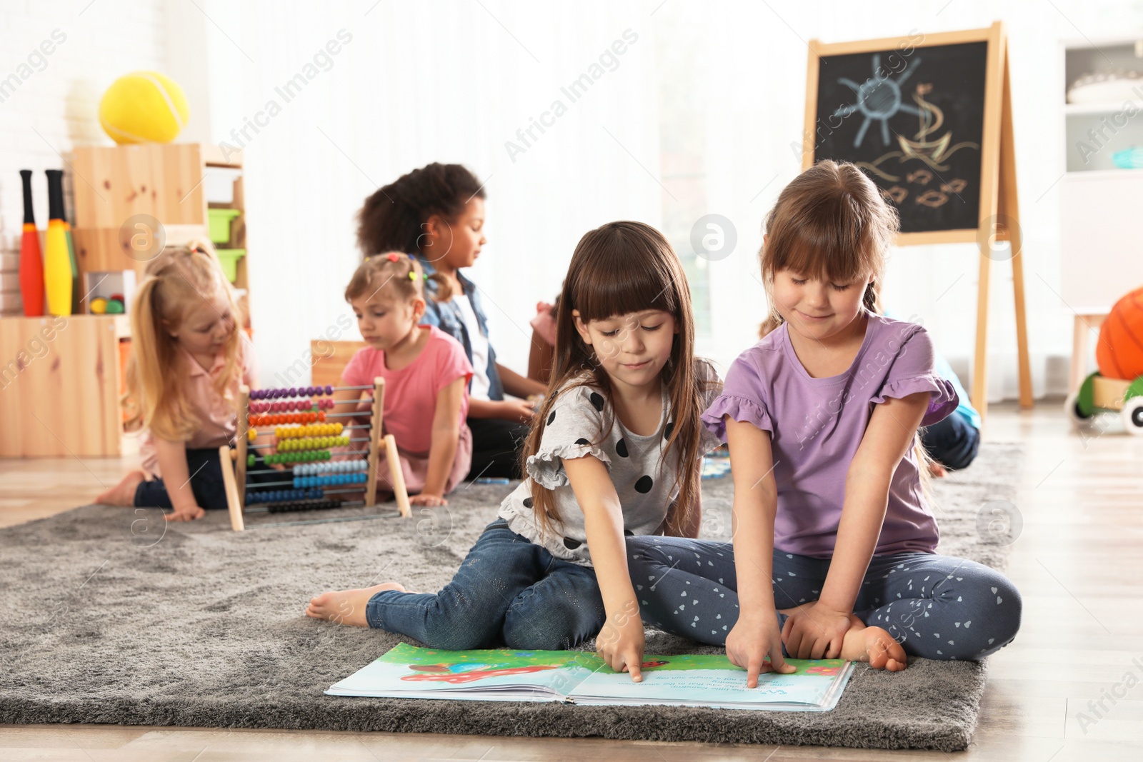 Photo of Cute girls reading book on floor while other children playing together in kindergarten