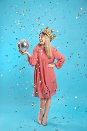 Happy young woman in party crown with disco ball and confetti on light blue background