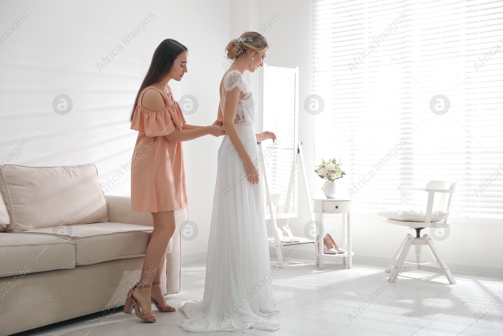 Photo of Young woman helping bride to put on wedding dress in room. Space for text