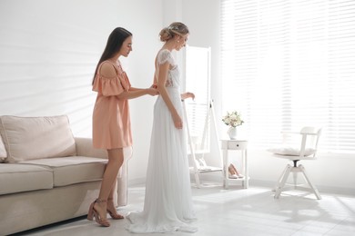 Young woman helping bride to put on wedding dress in room. Space for text