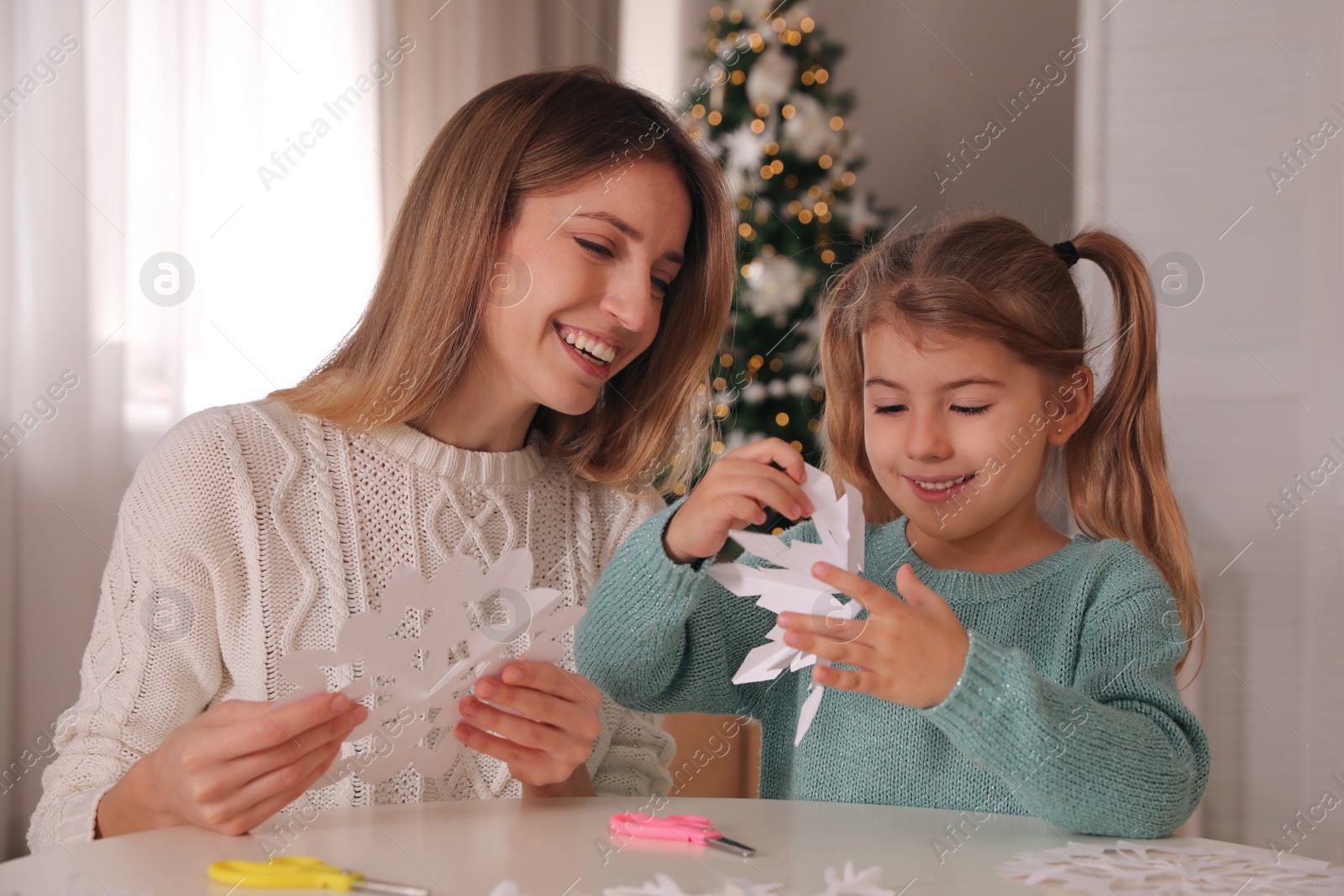 Photo of Happy mother and daughter making paper snowflakes at table near Christmas tree indoors