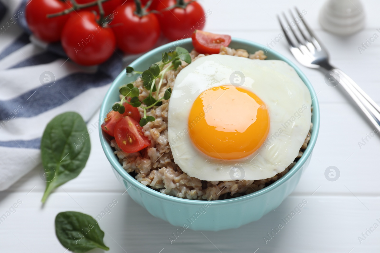 Photo of Tasty boiled oatmeal with fried egg, tomato and microgreens served on white wooden table, closeup