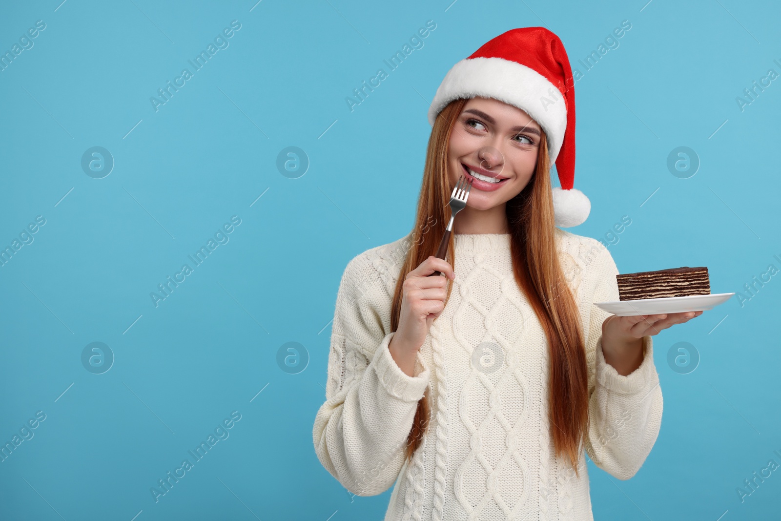 Photo of Young woman in Santa hat with piece of tasty cake on light blue background, space for text