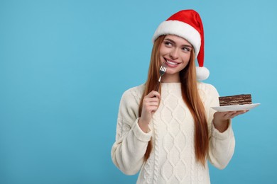 Young woman in Santa hat with piece of tasty cake on light blue background, space for text