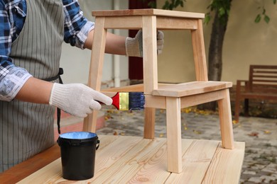 Photo of Man varnishing wooden step stool at table outdoors, closeup