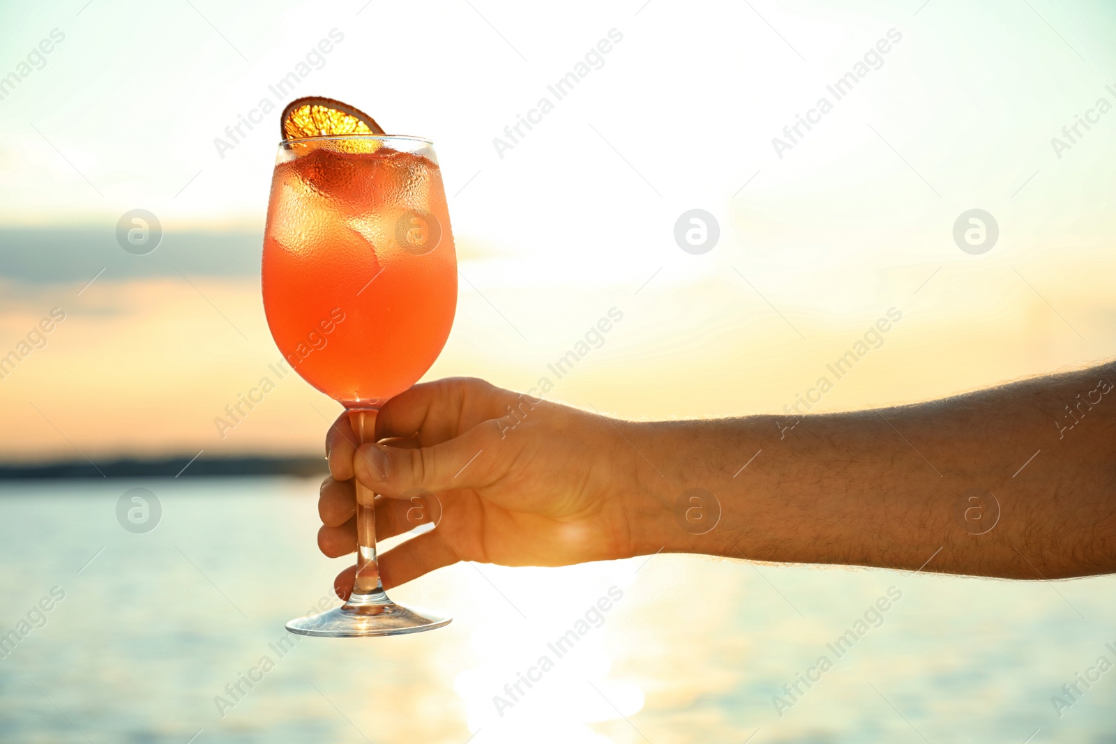 Photo of Man with glass of fresh summer cocktail near water at sunset, closeup