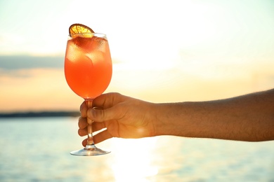 Man with glass of fresh summer cocktail near water at sunset, closeup