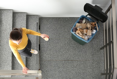 Photo of Young woman throwing coffee cup in trash bin indoors, top view. Waste recycling