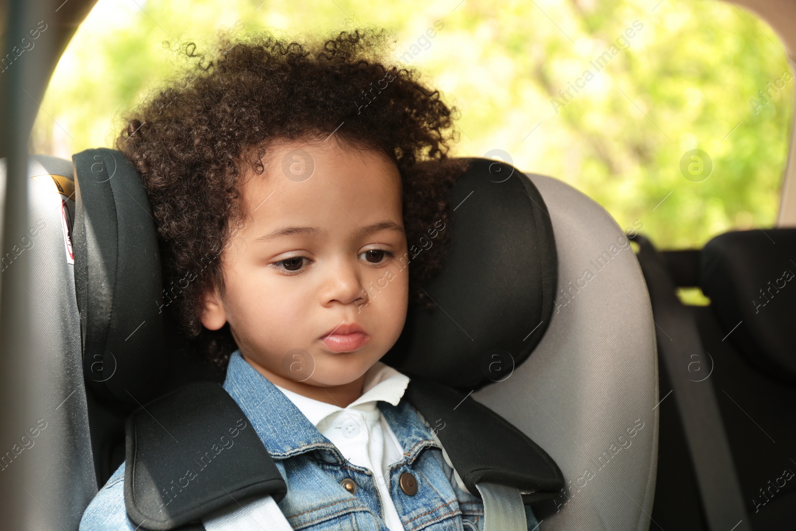 Photo of Cute African-American girl sitting in safety seat alone inside car. Child in danger