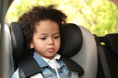 Photo of Cute African-American girl sitting in safety seat alone inside car. Child in danger