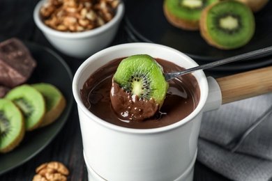 Photo of Dipping slice of kiwi into fondue pot with milk chocolate on black table, closeup