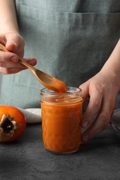 Woman taking delicious persimmon jam with spoon from glass jar at gray table, closeup