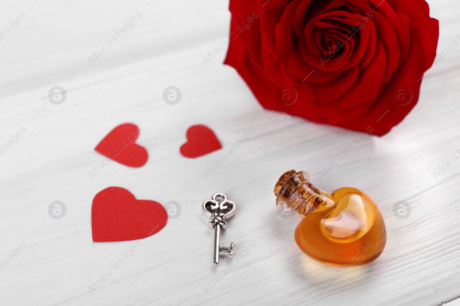 Photo of Heart shaped bottle of love potion with small key, paper hearts and red rose flower on white wooden table