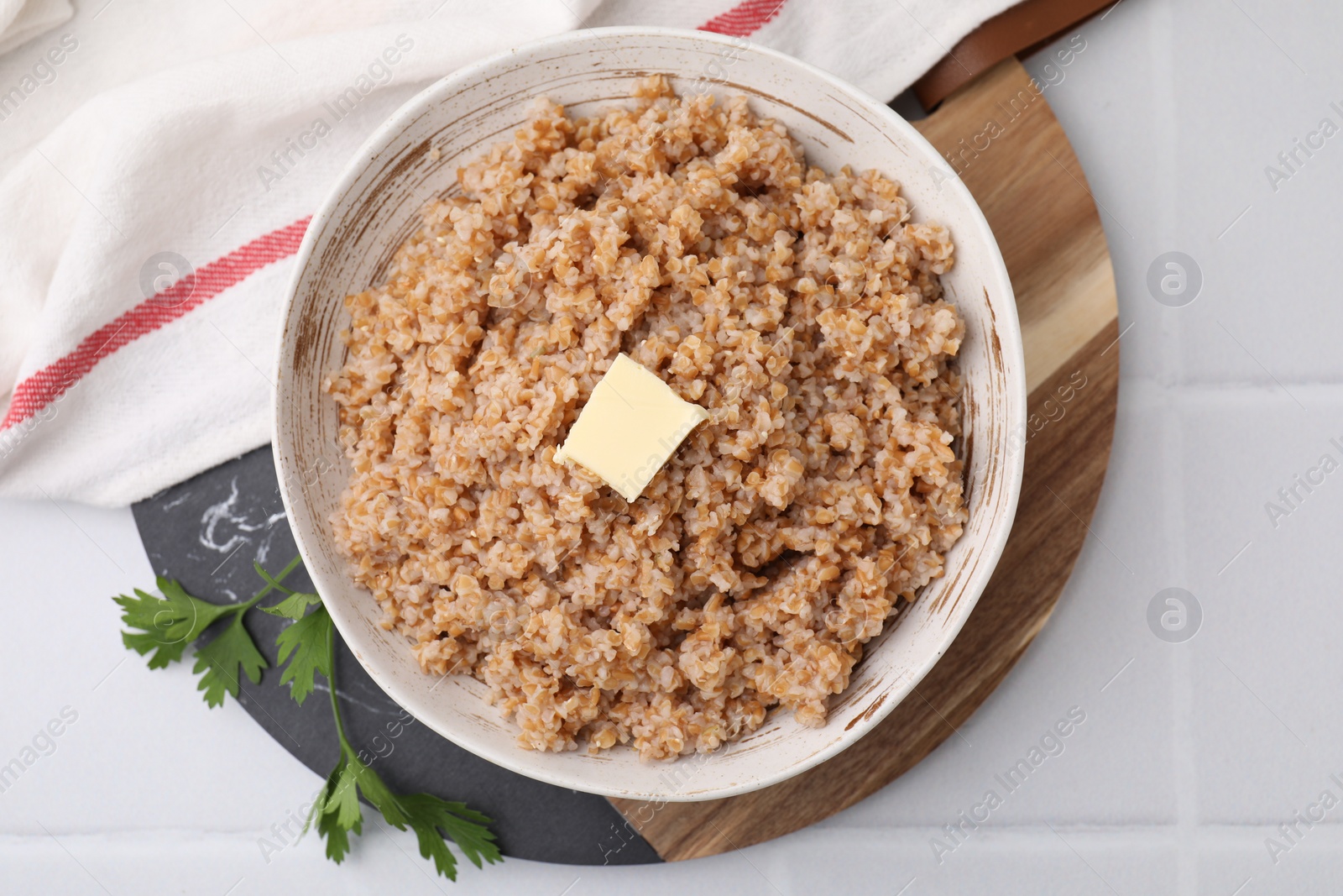 Photo of Tasty wheat porridge with butter and parsley in bowl on white tiled table, top view