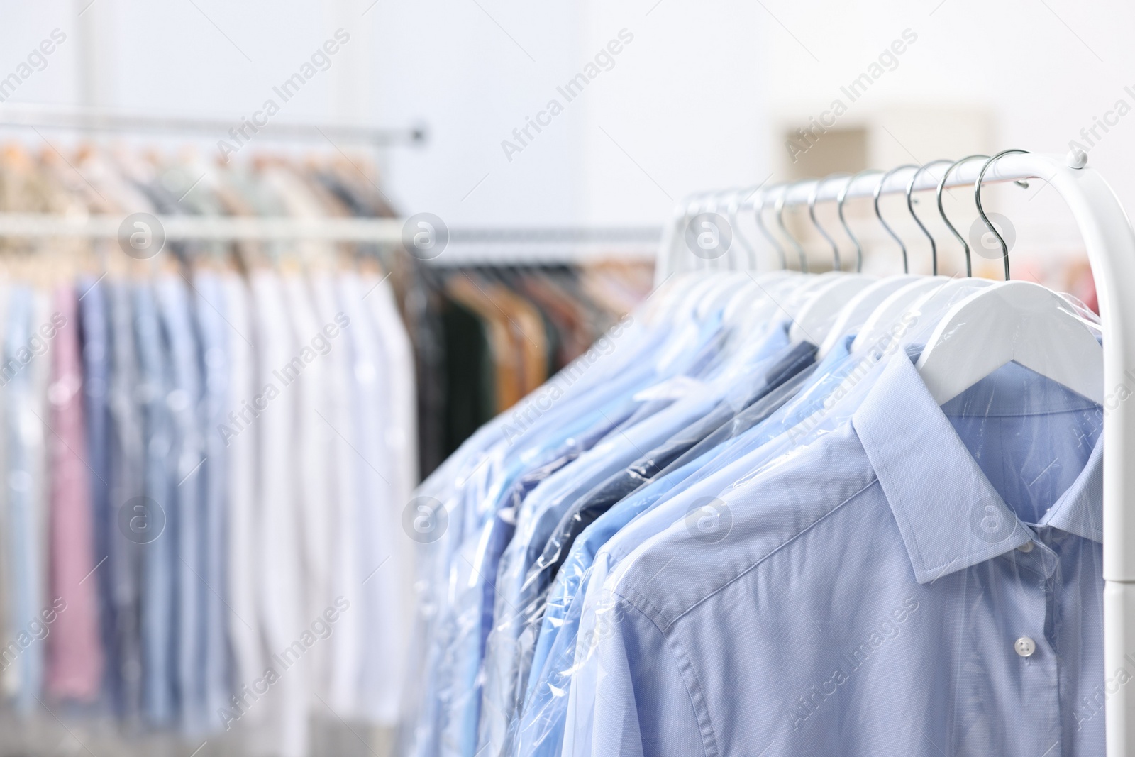 Photo of Dry-cleaning service. Many different clothes in plastic bags hanging on rack indoors, closeup