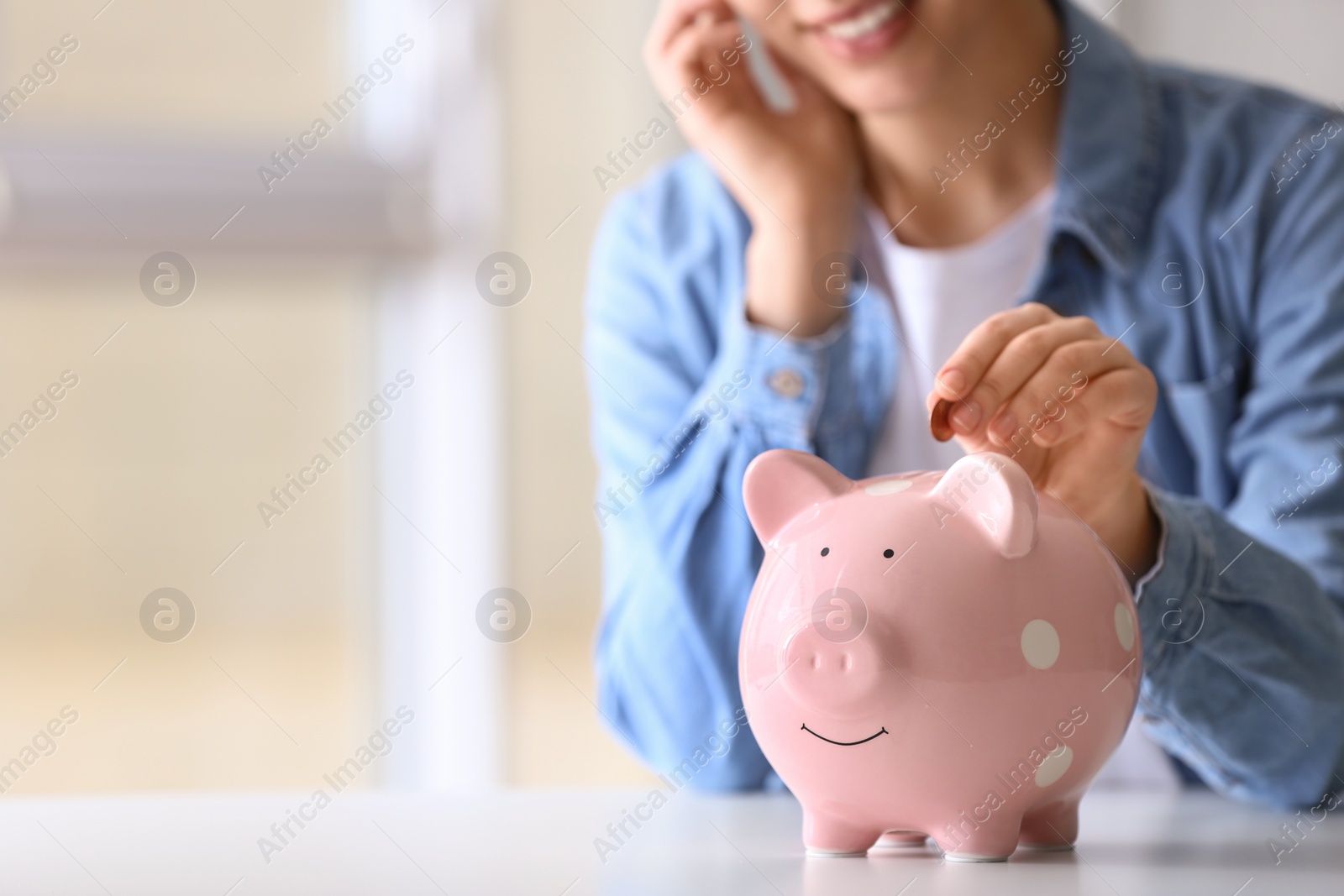 Photo of Woman putting coin into piggy bank at table indoors, closeup. Space for text